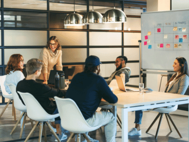 Group of six people in a modern office engaged in a meeting. A woman stands by a whiteboard with sticky notes. They appear focused and collaborative.