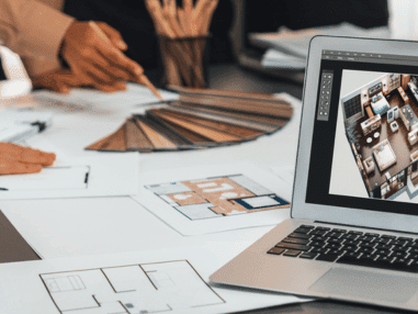 Laptop displaying a detailed floor plan, surrounded by printed blueprints and color swatches. A hand points to the plans, suggesting collaborative design work.