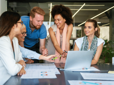 A diverse group of five colleagues, three women and two men, collaborate around a table with laptops and documents, smiling and engaged in discussion.