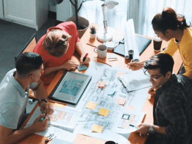 A diverse group of four people collaboratively reviewing architectural drawings at a table, surrounded by digital devices, notes, and coffee, conveying teamwork.