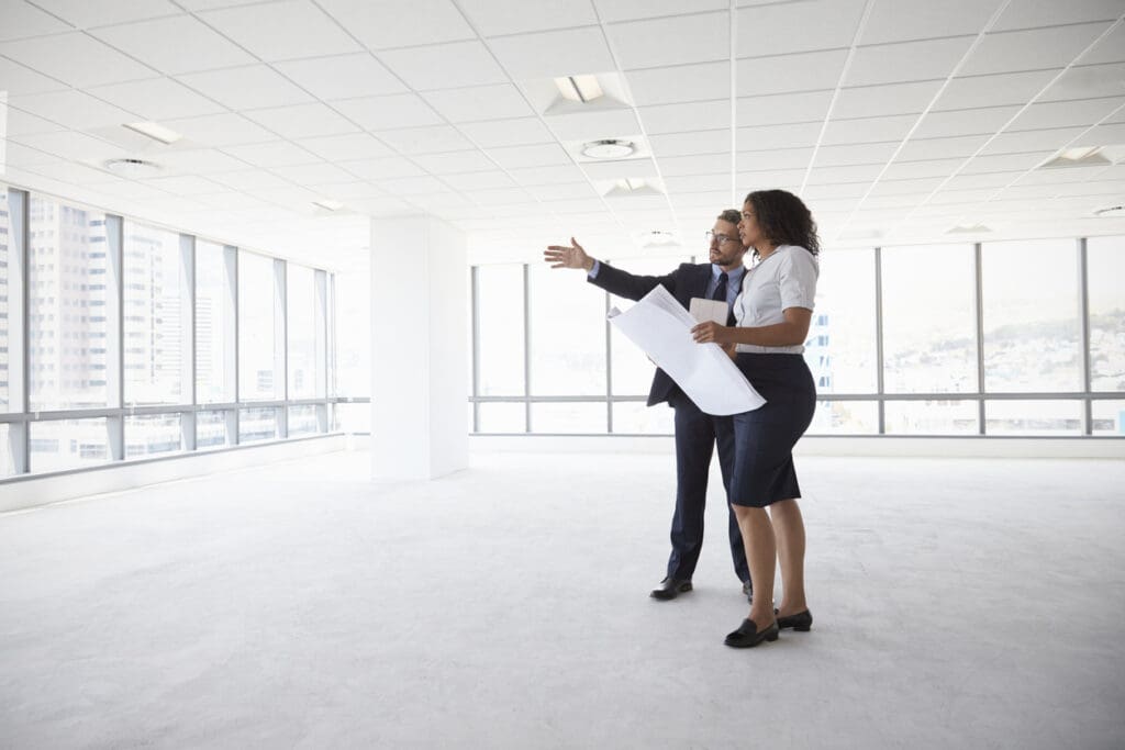 Two people reviewing floor plans in a bright empty commercial office space with large windows, suspended ceiling tiles and open floor area, showing the planning stage of a Cat A fit out.
