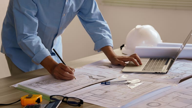 Person reviewing architectural floor plans and working on a laptop beside construction drawings, measuring tools and a hard hat, representing end to end project management for an office design and fit out project.