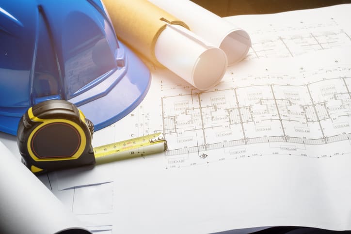 Blue construction hard hat, tape measure and rolled architectural plans laid across detailed office floor plan drawings for an office refurbishment project.