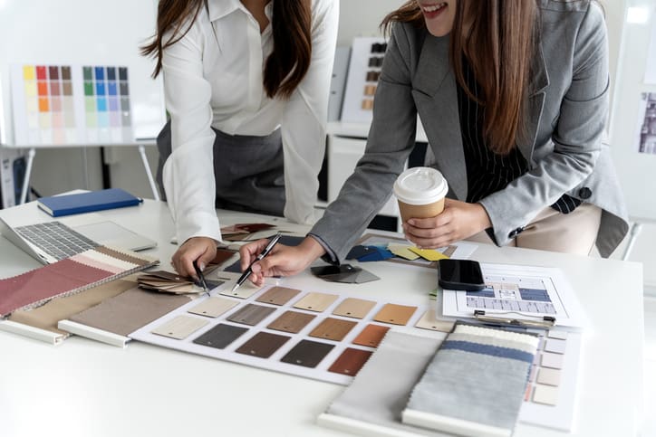 Two interior designers reviewing colour swatches, fabric samples, material finishes and floor plans at a design table while developing an office interior scheme.