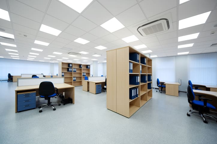 Bright open-plan office interior with individual desks, blue and black task chairs, wooden storage shelving, filing cabinets, suspended ceiling panels and integrated lighting, representing a completed office refurbs project.